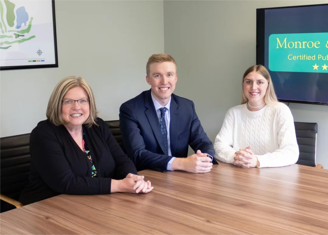 Three people sit at a conference table, smiling. A sign in the background reads "Monroe CPA".