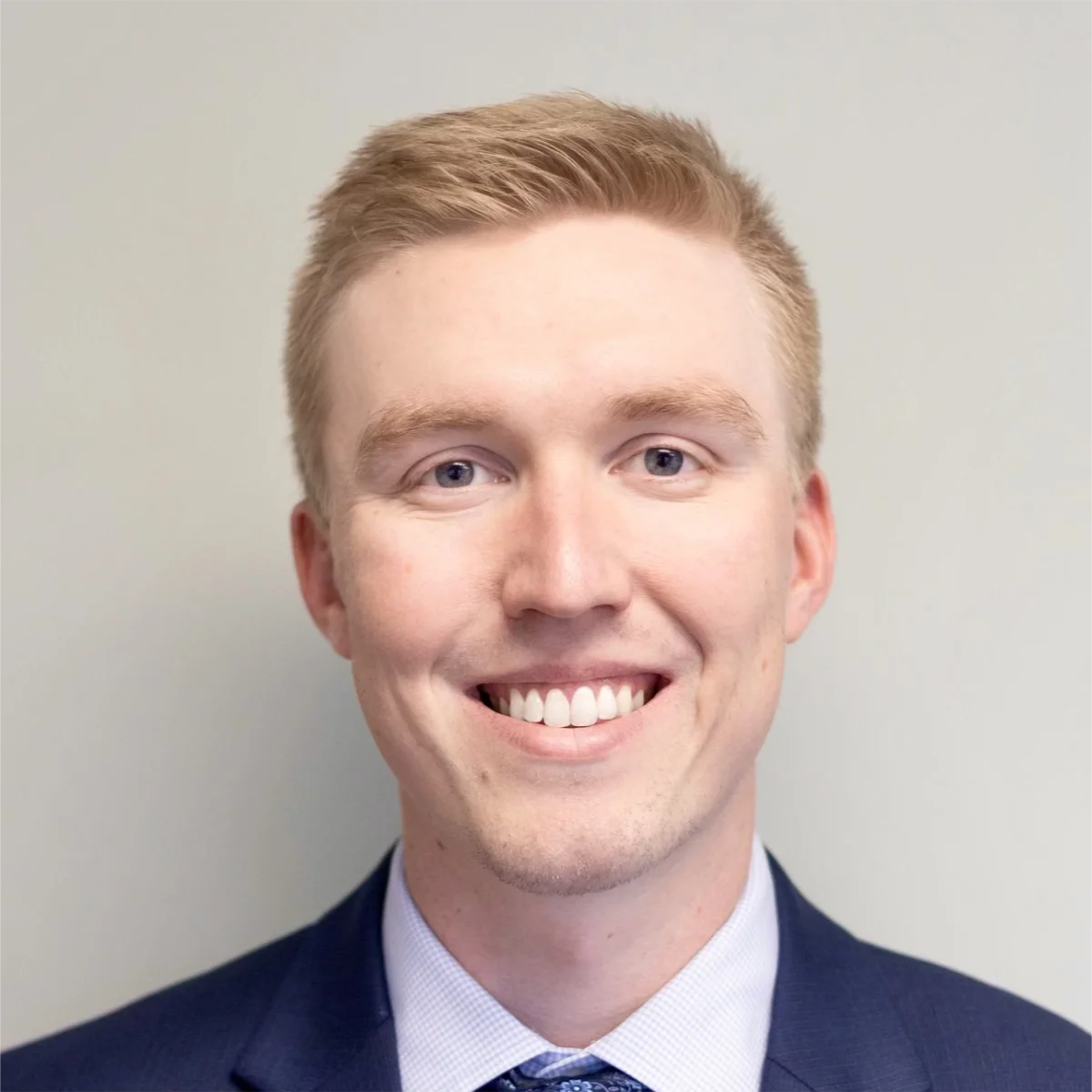 A person with short blond hair smiles, wearing a navy suit jacket and blue tie in front of a plain background.